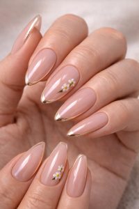 Glossy nude almond nails with thin gold outline tips and tiny white daisy flower accents, elegant Nail Trends 2026 at Top Nails 3 Airdrie